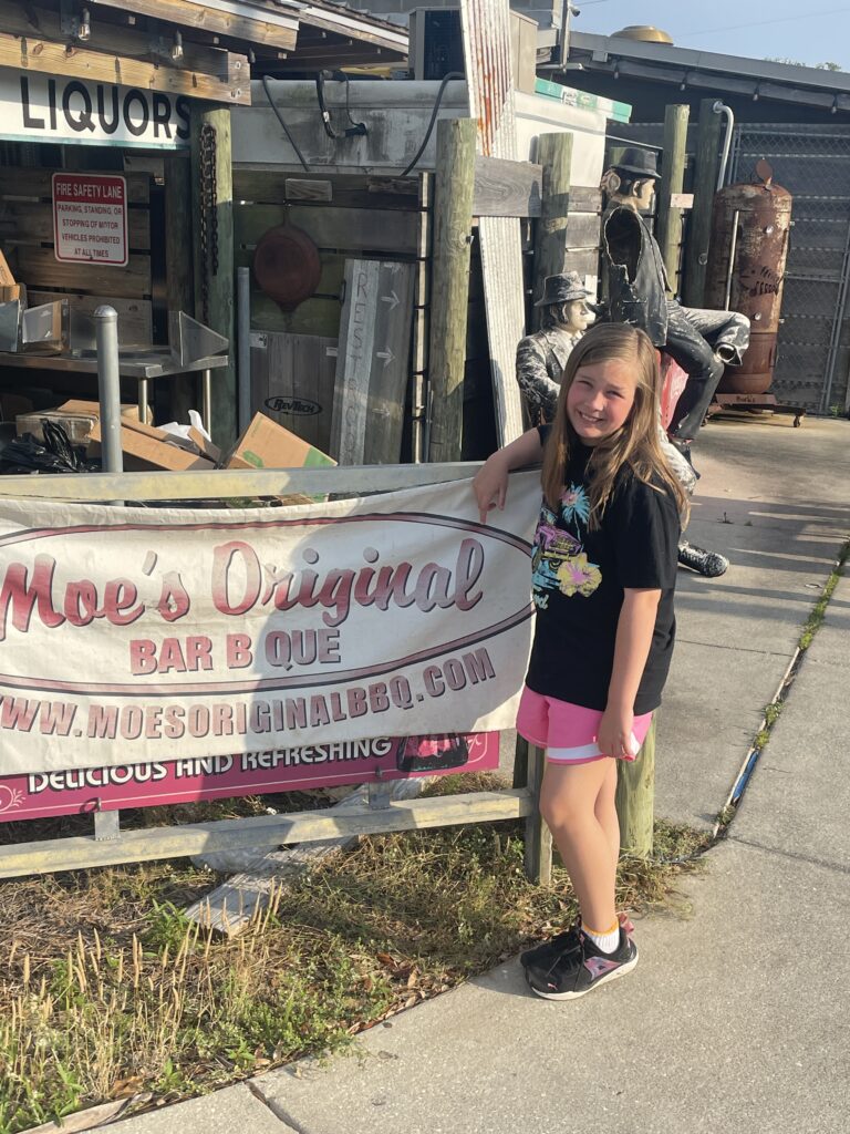 Young girl standing beside a sign that reads Moe's Original BBQ