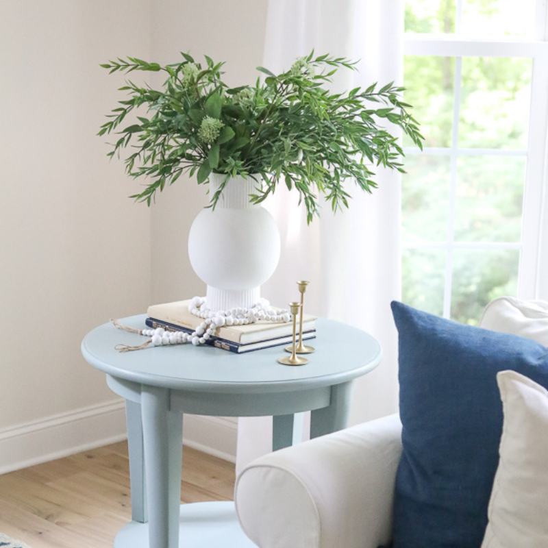 light blue end table with white planter and green plant sitting on books. the table is beside a white couch with a navy blue pillow on it.
