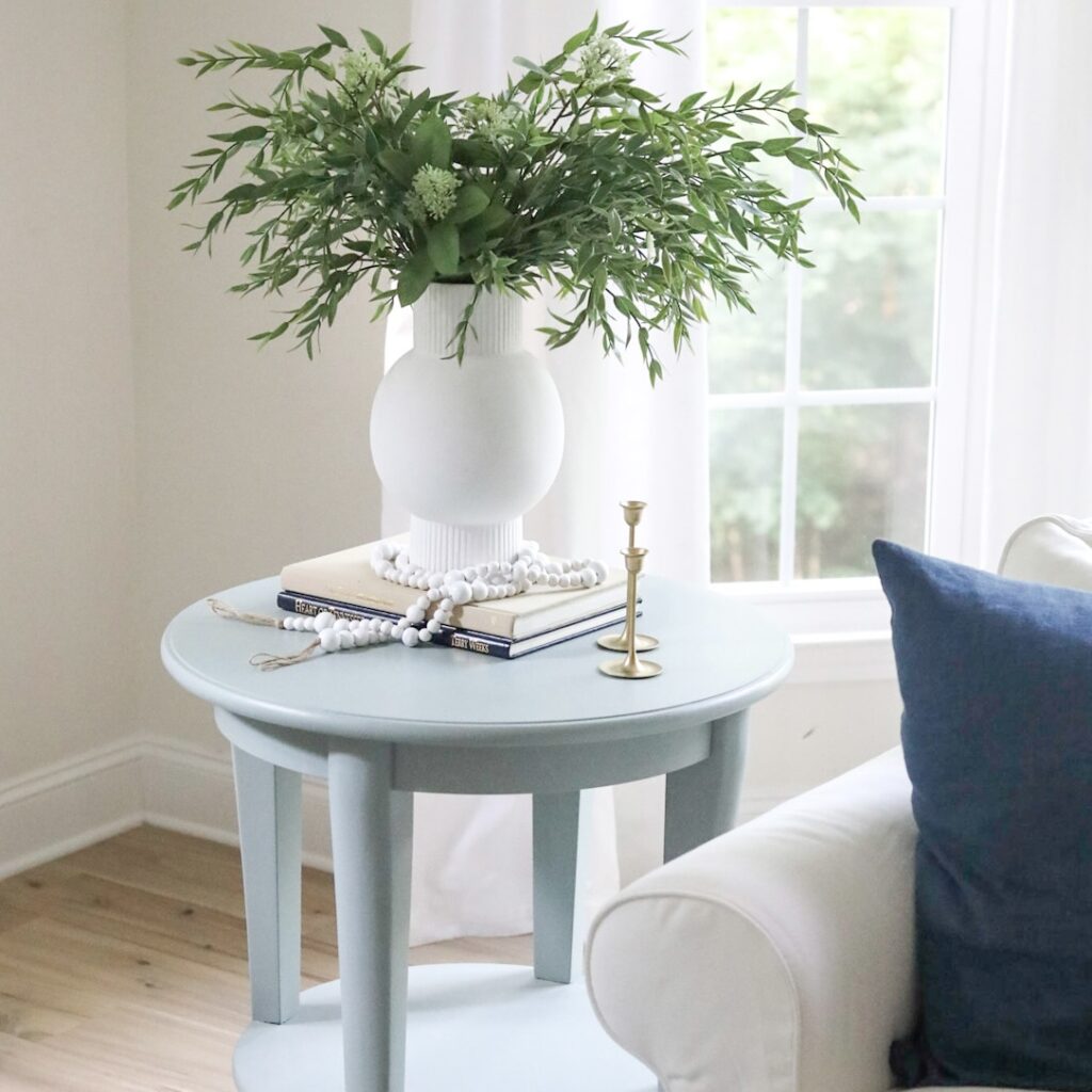 round side table with white vase and flowery greenery beside a white couch with blue accent pillow